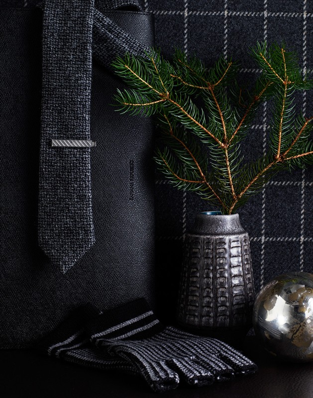 Still life of a dark gray tie with a tie clip, gloves, an ornamental ball, and a vase with pine branches on a plaid background.