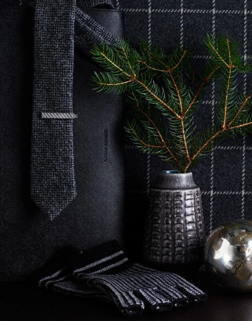 Still life of a dark gray tie with a tie clip, gloves, an ornamental ball, and a vase with pine branches on a plaid background.