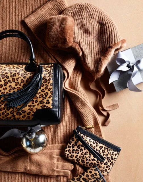 Cozy winter accessories flat lay: leopard print handbag, knitted hat, gloves, sweater, gift box on tan backdrop for fashion editorial.