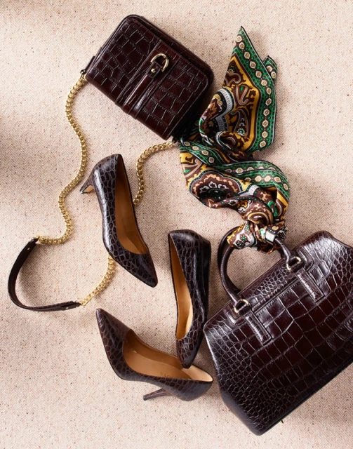 Flat lay composition of a brown alligator purse, handbag, pumps, and a patterned scarf on a light-colored textured surface.