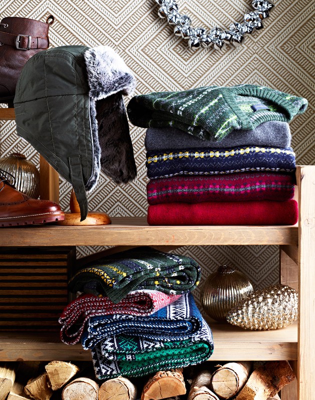 Still life composition featuring a wood shelf with stacked sweaters, a hat with ear flaps, leather boots and shoes, and metal orbs.