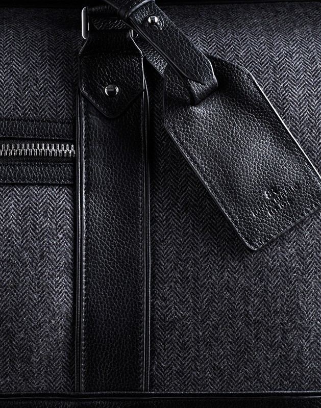 Close-up shot of a stylish gray herringbone-patterned duffel bag with black leather accents and a zipper, suitable for travel or fashion.