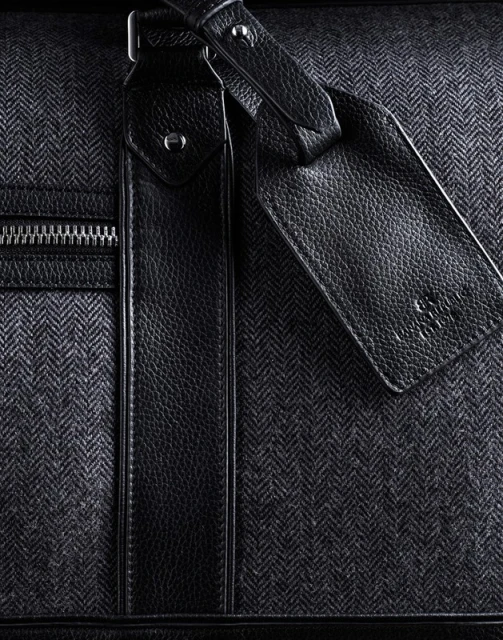 Close-up shot of a stylish gray herringbone-patterned duffel bag with black leather accents and a zipper, suitable for travel or fashion.