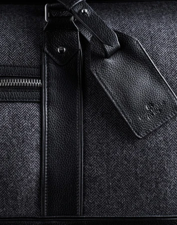 Close-up shot of a stylish gray herringbone-patterned duffel bag with black leather accents and a zipper, suitable for travel or fashion.