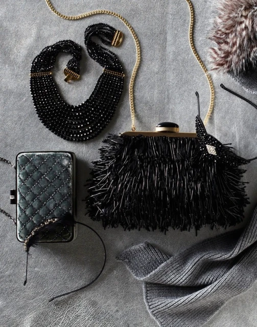 Black beaded necklace and fringe purse with chain strap are staged with a quilted wallet and gray wool scarf on a gray background.