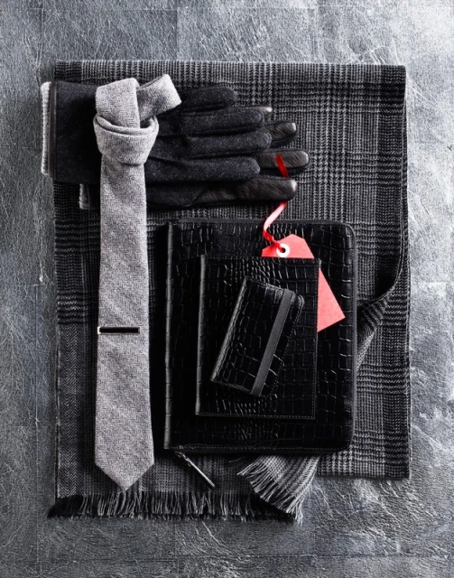 Still life of grey necktie, black leather gloves, planner, and cardholder, displayed on a grey wool scarf and surface.