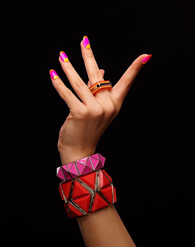 Hand with pink and yellow manicured nails wearing a ring and two bracelets in red and pink against a dark black background.