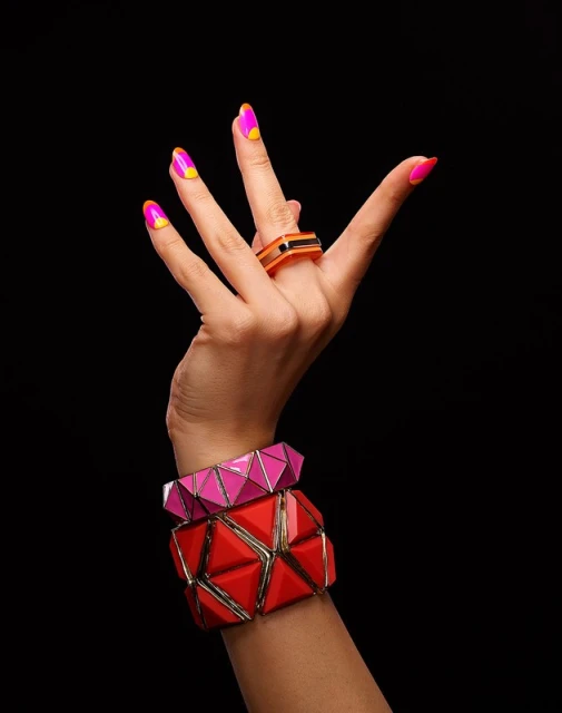 Hand with pink and yellow manicured nails wearing a ring and two bracelets in red and pink against a dark black background.