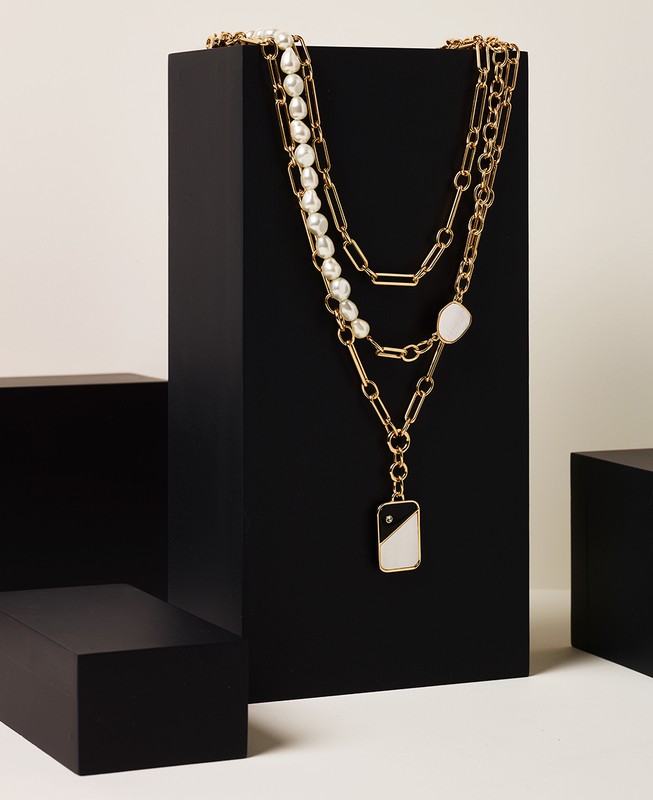 Three layered necklaces on a tiered black display: a pearl necklace, a gold chain, and a long pendant necklace against a white background.