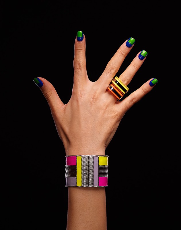Close-up of a hand featuring blue and green manicured nails and gold jewelry, including a chunky ring and geometric bracelet.