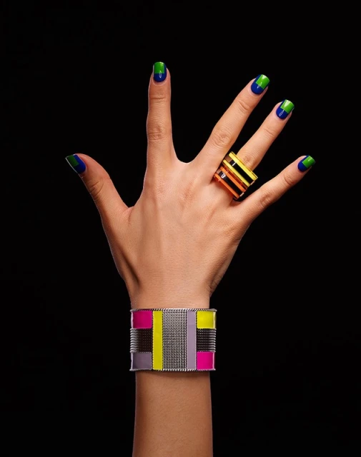 Close-up of a hand featuring blue and green manicured nails and gold jewelry, including a chunky ring and geometric bracelet.