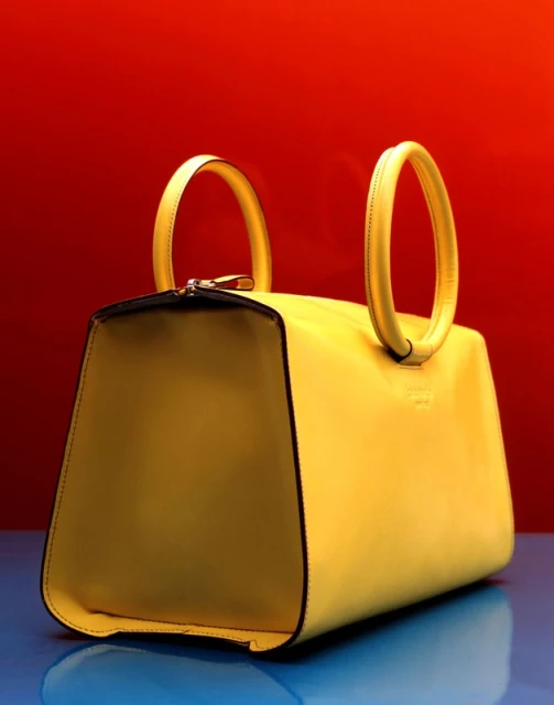Close-up studio shot of a yellow leather handbag with black trim and round handles against a red and blue gradient background.