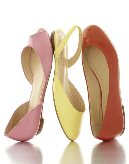 Close-up shot of three colorful patent leather ballet flats in pink, yellow, and red, arranged on a white surface with a bright background.