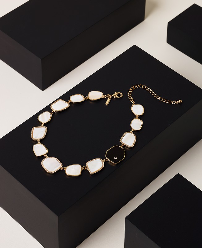 Gold necklace with off-white and black stone accents displayed on black cubes against an off-white backdrop in a studio setting.