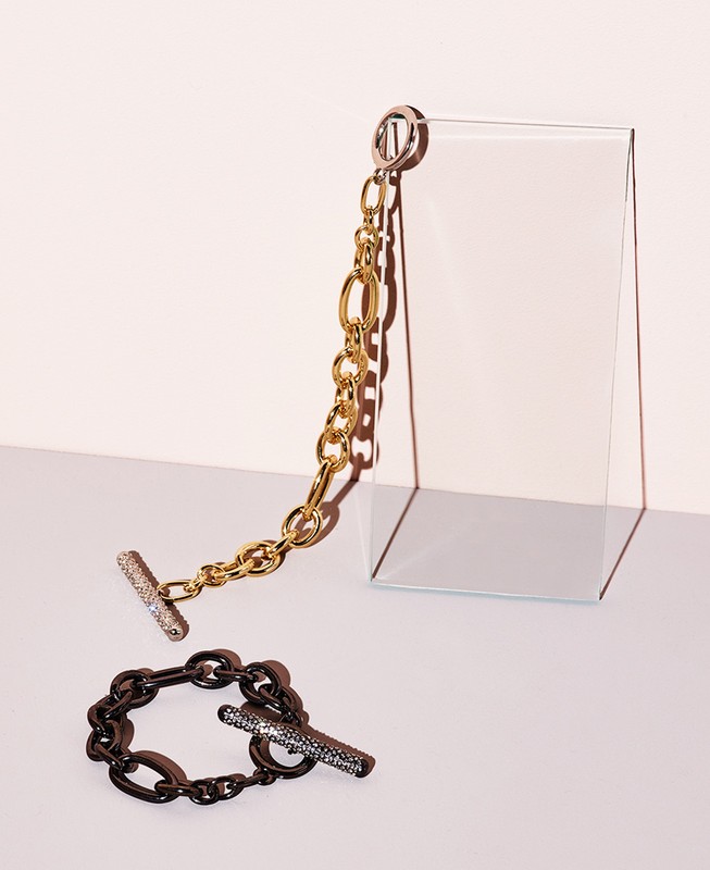 Close-up studio shot of two fashion bracelets; one gold chain link and one black chain link on a light pink background with a glass prop.