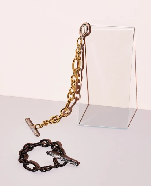 Close-up studio shot of two fashion bracelets; one gold chain link and one black chain link on a light pink background with a glass prop.
