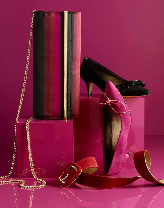 Elegant fashion product shot: a clutch with a gold chain, black and pink heels, and a red velvet belt on a pink cube against a pink backdrop.