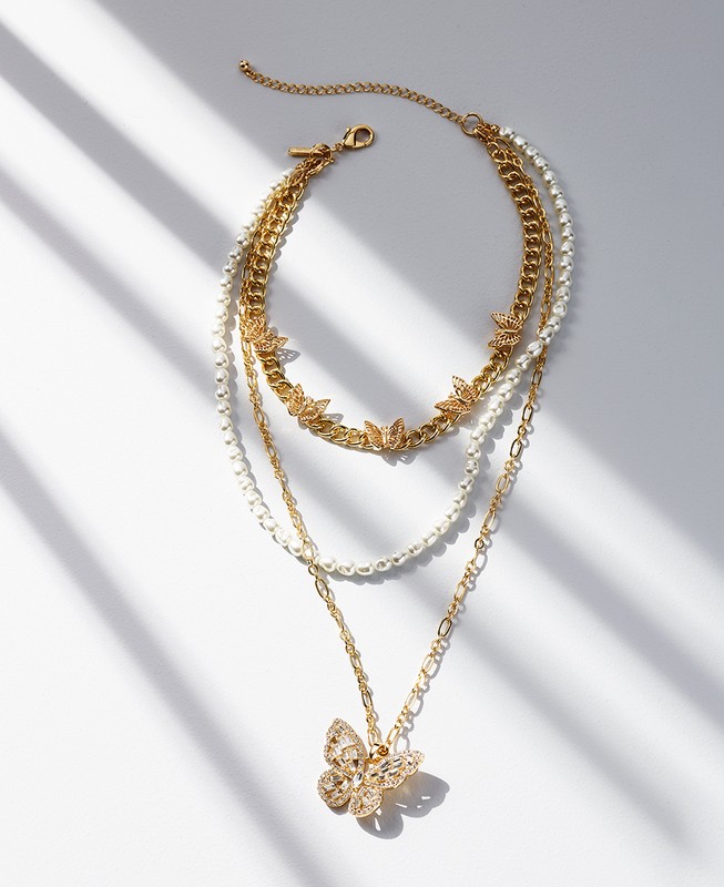 A layered gold chain necklace featuring a butterfly pendant, showcased on a white surface with sunlight creating soft shadows.
