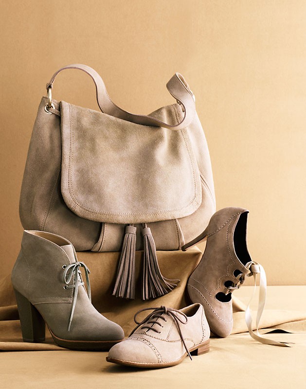 Still life studio shot of a taupe-colored suede shoulder bag, ankle boots with block heels, a pair of oxfords, and a high-heeled shoe.