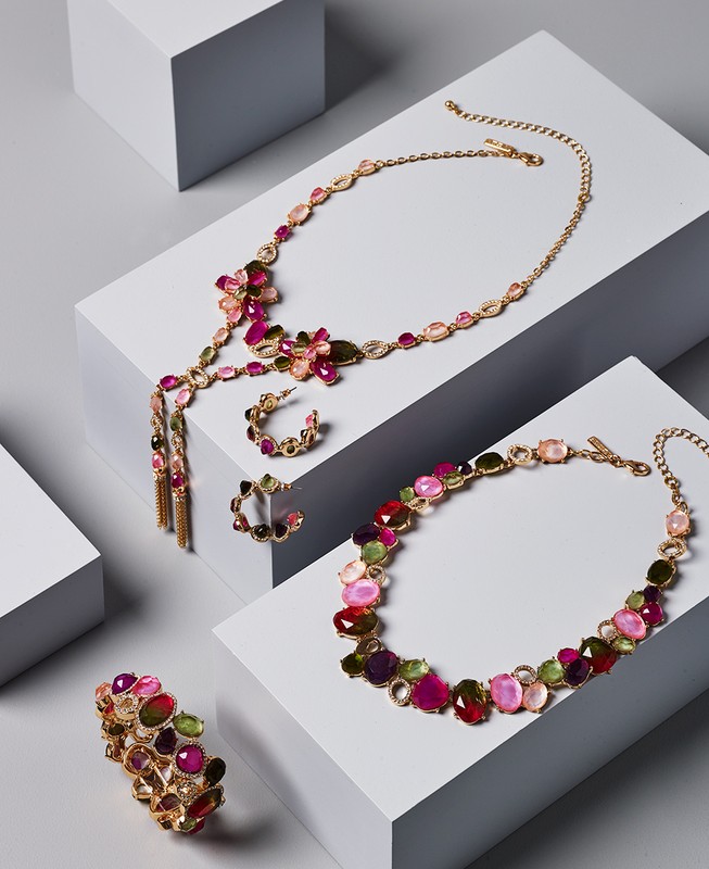 Still life of four gold jewelry pieces with pink, green, and clear stones arranged on white geometric platforms against a gray backdrop.
