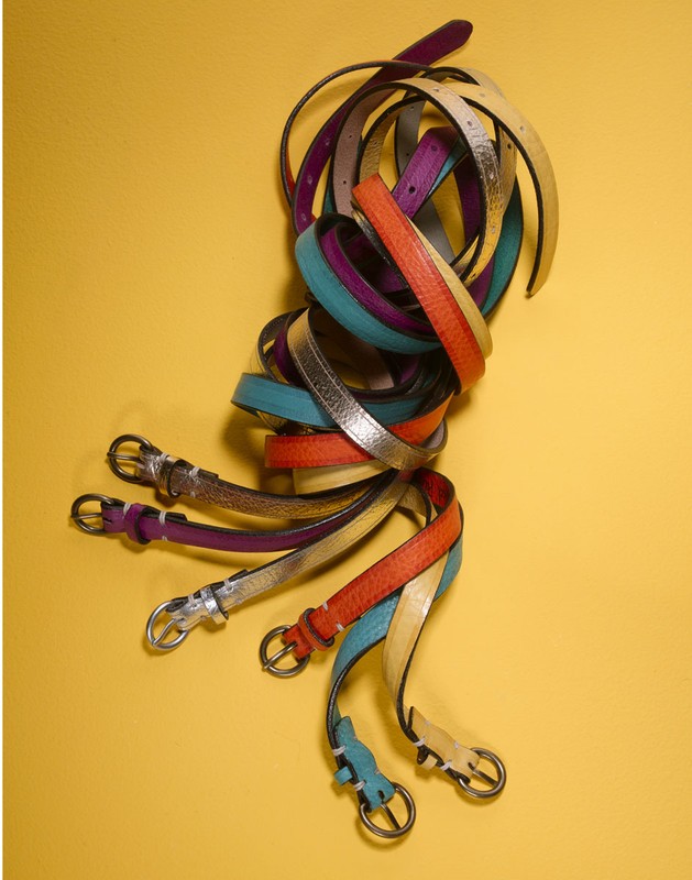 Fashion belts of purple, teal, gold, and orange are coiled together on a yellow background, creating a bright, stylish composition.