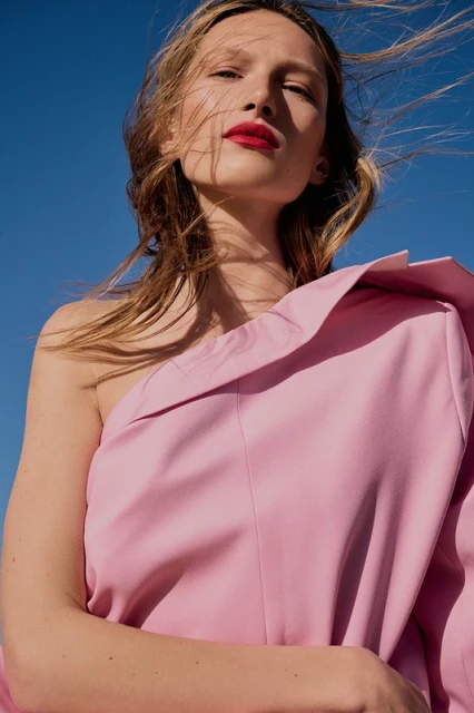 Blonde woman poses in a pink one-shoulder dress against a clear blue sky, highlighting fashion and beauty in an outdoor setting.