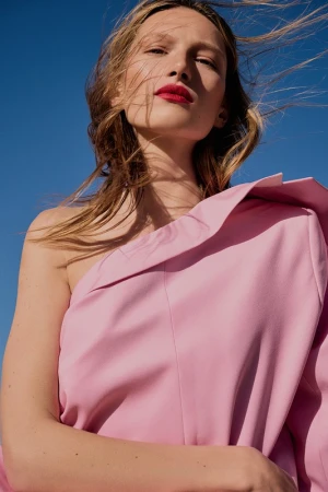 Blonde woman poses in a pink one-shoulder dress against a clear blue sky, highlighting fashion and beauty in an outdoor setting.