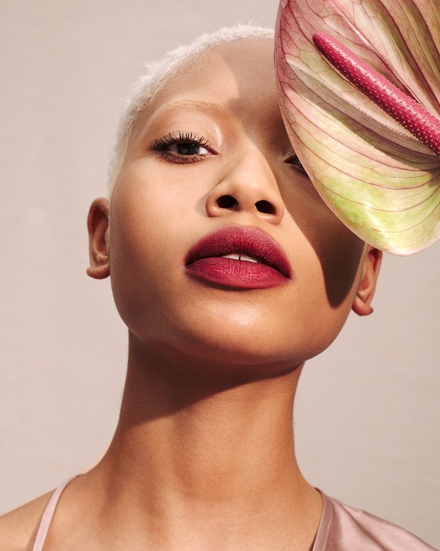 Close-up of a fair-skinned adult woman with short white hair and dark red lipstick, posing with a pink anthurium flower.