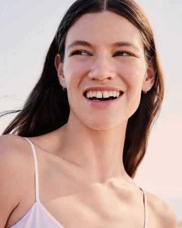 Close-up portrait of a smiling young woman with dark hair wearing a light-coloured strap top against a clear sky background.
