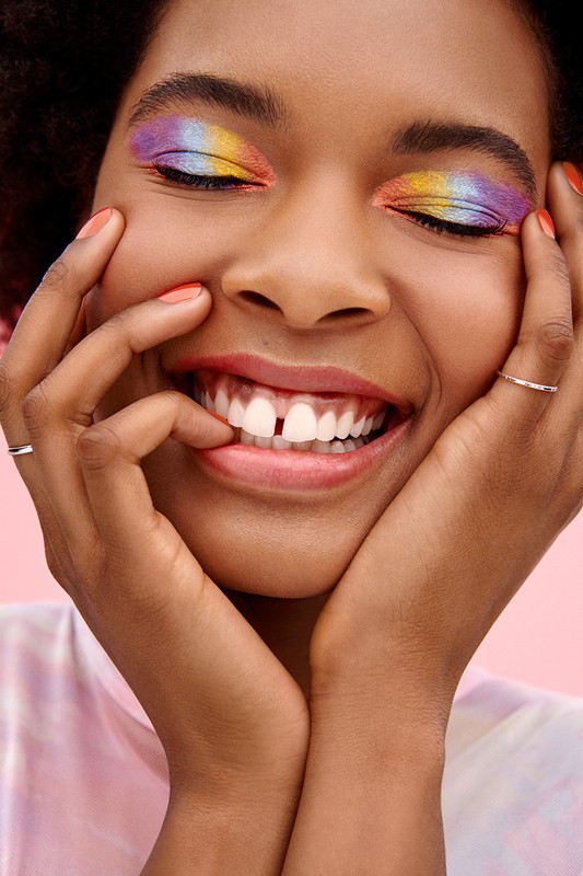 Smiling Black woman with rainbow eyeshadow holds face in hands, showcasing vibrant makeup against a soft pink backdrop.