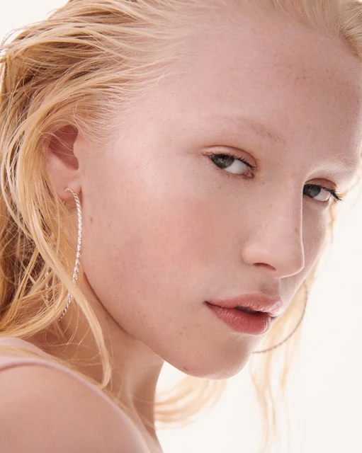 Close-up portrait of a fair-skinned woman with blonde hair wearing hoop earrings, set against a white backdrop in a beauty-style image.