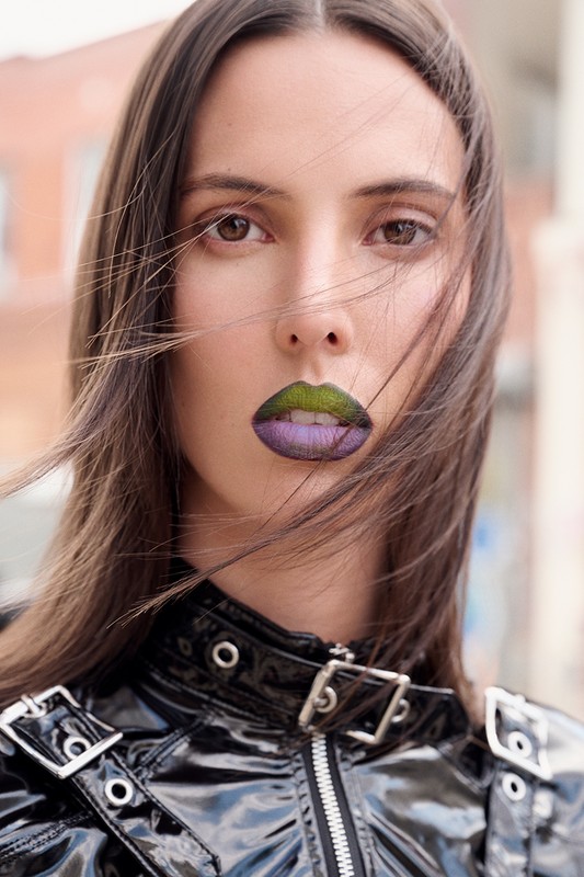 Close-up of a woman with long brown hair, wearing a black vinyl jacket with silver hardware, and a unique green and purple lip color.