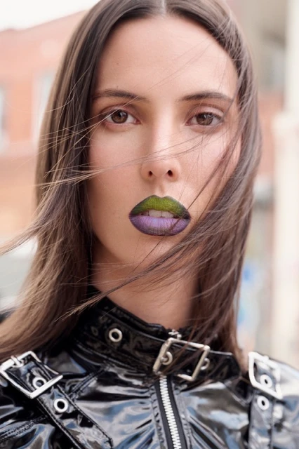 Close-up of a woman with long brown hair, wearing a black vinyl jacket with silver hardware, and a unique green and purple lip color.
