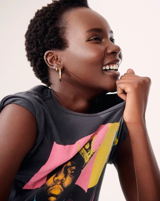 Woman smiles and wears a graphic tee shirt with image of The Notorious B.I.G. in a bright, neutral studio setting.