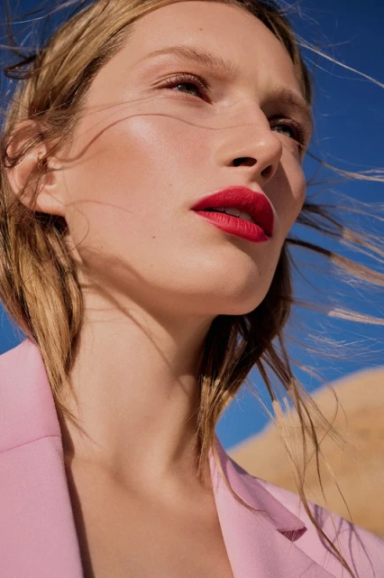 Woman with blonde hair and bright red lips wearing a pink jacket, set against a backdrop of clear blue sky and desert landscape.