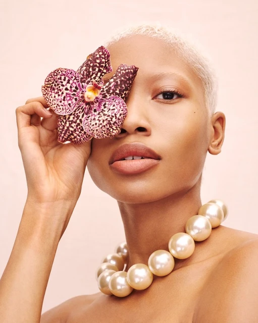 Close-up of a fair-skinned woman with a platinum blonde buzzcut, wearing a pearl necklace and holding a speckled orchid to her face.