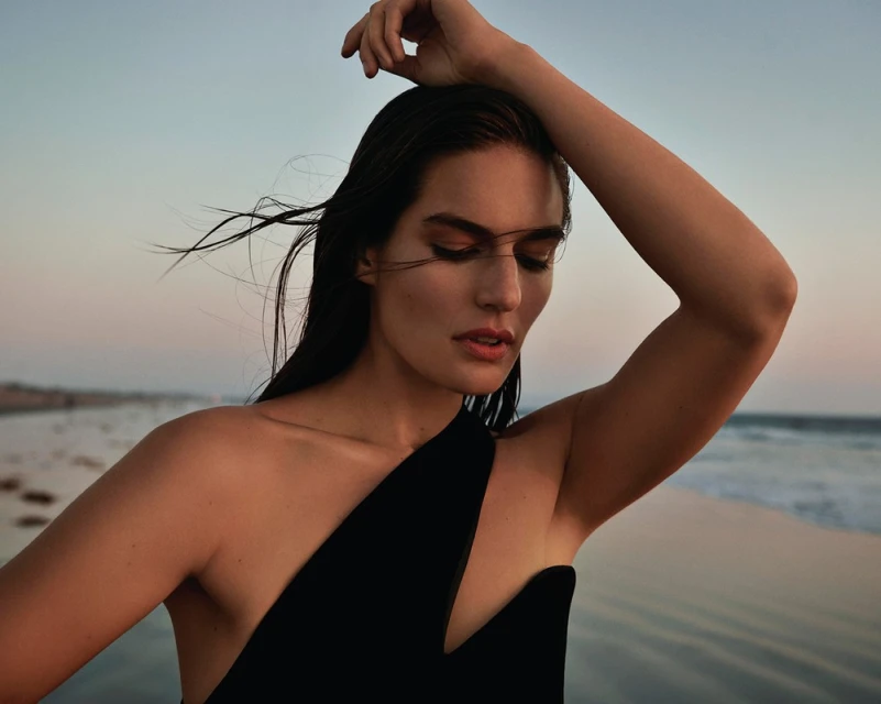 Adult female portrait on a beach at sunset wearing a black dress. The model is centered with natural light highlighting wet hair.
