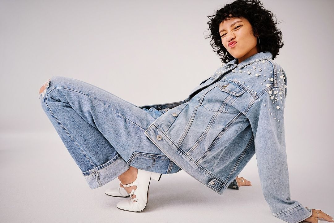 A young woman with curly dark hair wearing a denim jacket with pearls, ripped jeans, and white ballet flats, posing in a studio.