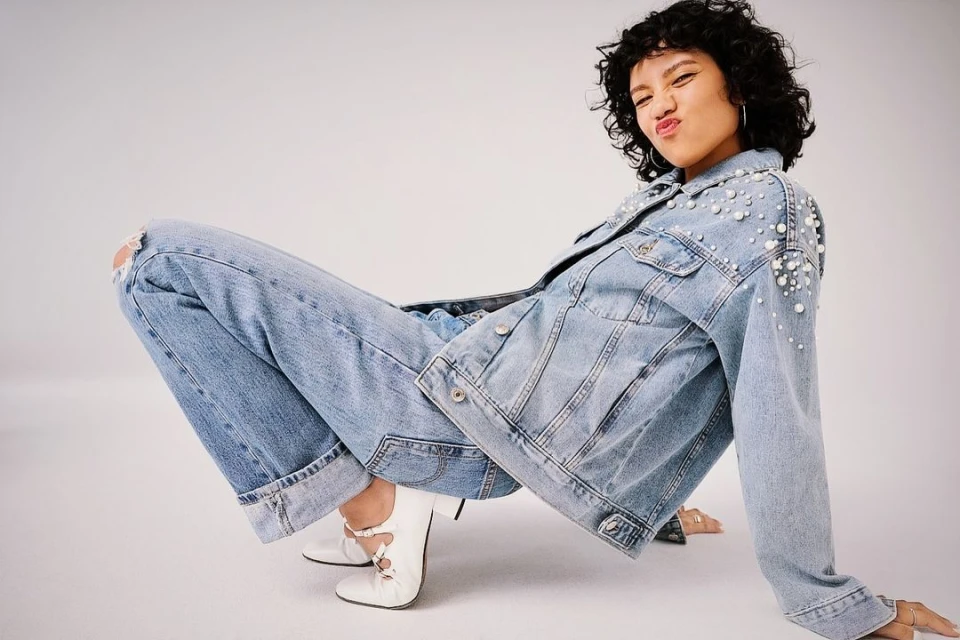 A young woman with curly dark hair wearing a denim jacket with pearls, ripped jeans, and white ballet flats, posing in a studio.