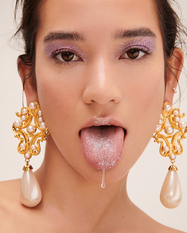 Close-up portrait of a woman with glitter eyelids and tongue sticking out wearing ornate pearl drop earrings.