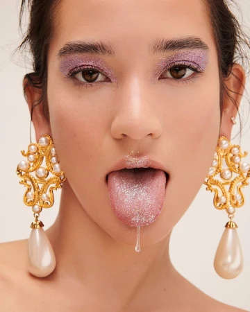 Close-up portrait of a woman with glitter eyelids and tongue sticking out wearing ornate pearl drop earrings.
