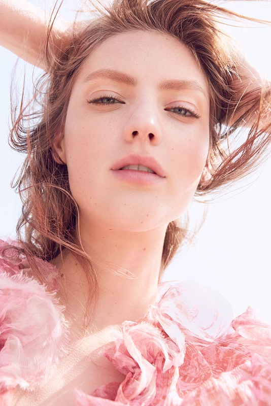 Close-up portrait of a young adult female with light brown hair, fair skin, and pink ruffled clothing against a bright, clear sky.
