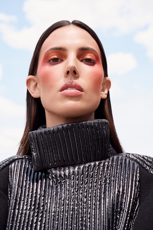 Woman with bold makeup wearing a high-neck black quilted jacket against a bright sky background. Beauty and fashion close-up.
