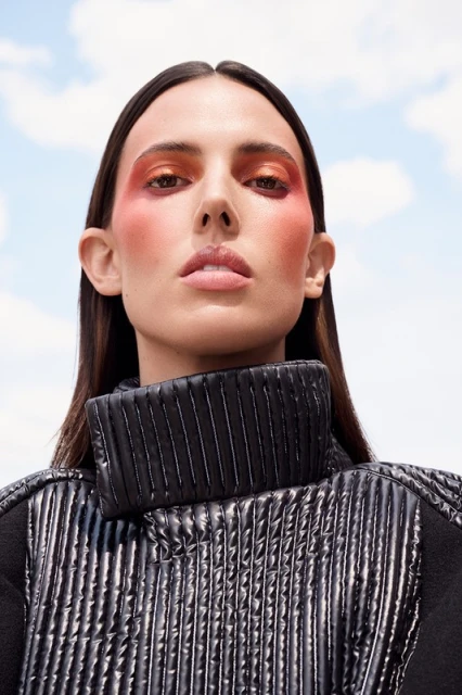 Woman with bold makeup wearing a high-neck black quilted jacket against a bright sky background. Beauty and fashion close-up.