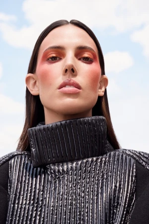 Woman with bold makeup wearing a high-neck black quilted jacket against a bright sky background. Beauty and fashion close-up.