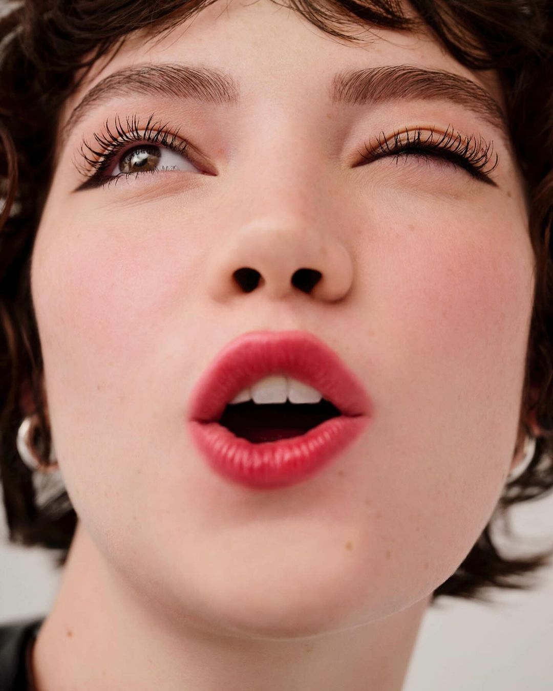 Close-up studio shot of a young adult female model with red lipstick, black eyeliner, and short brown hair winking at the camera.