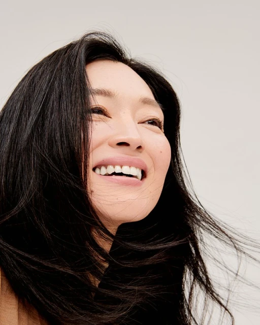 Close-up portrait of a smiling adult woman with dark hair, looking up against a light background, capturing her radiant expression and wind-swept hair.