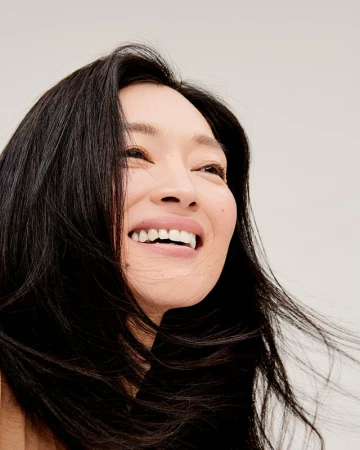 Close-up portrait of a smiling adult woman with dark hair, looking up against a light background, capturing her radiant expression and wind-swept hair.