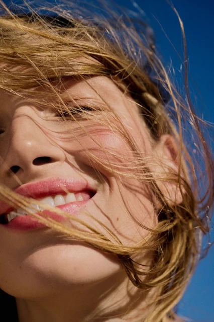 Close-up of a blonde woman smiling outdoors against a blue sky. The bright sunlight accentuates her features, and her hair is windswept.