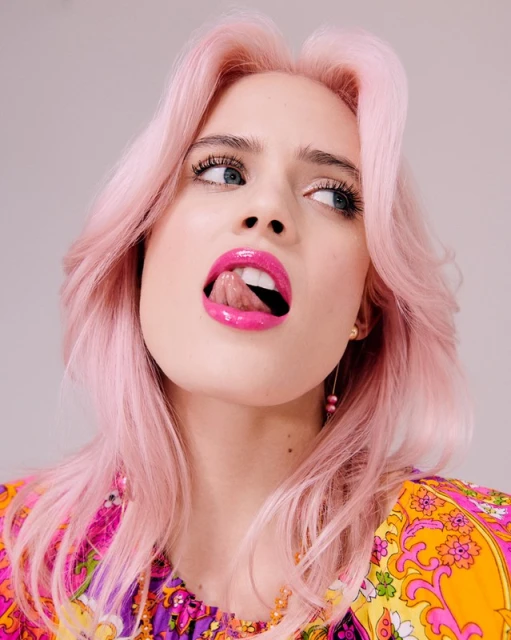 Close-up of a woman with pink hair wearing a colorful top sticking out her tongue, posing against a neutral studio backdrop.